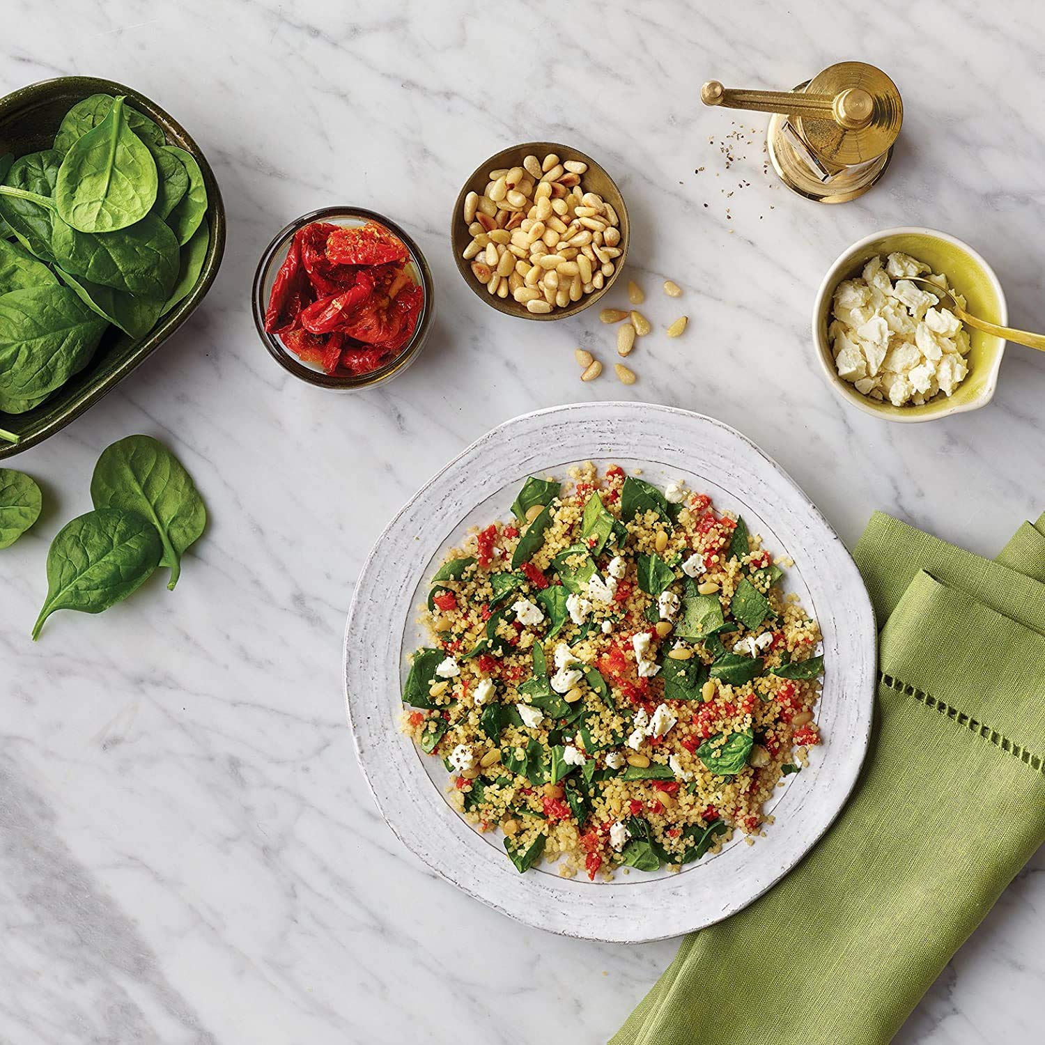 Mediterranean curry couscous served with spinach, sun-dried tomatoes, pine nuts, and feta cheese on a marble table.