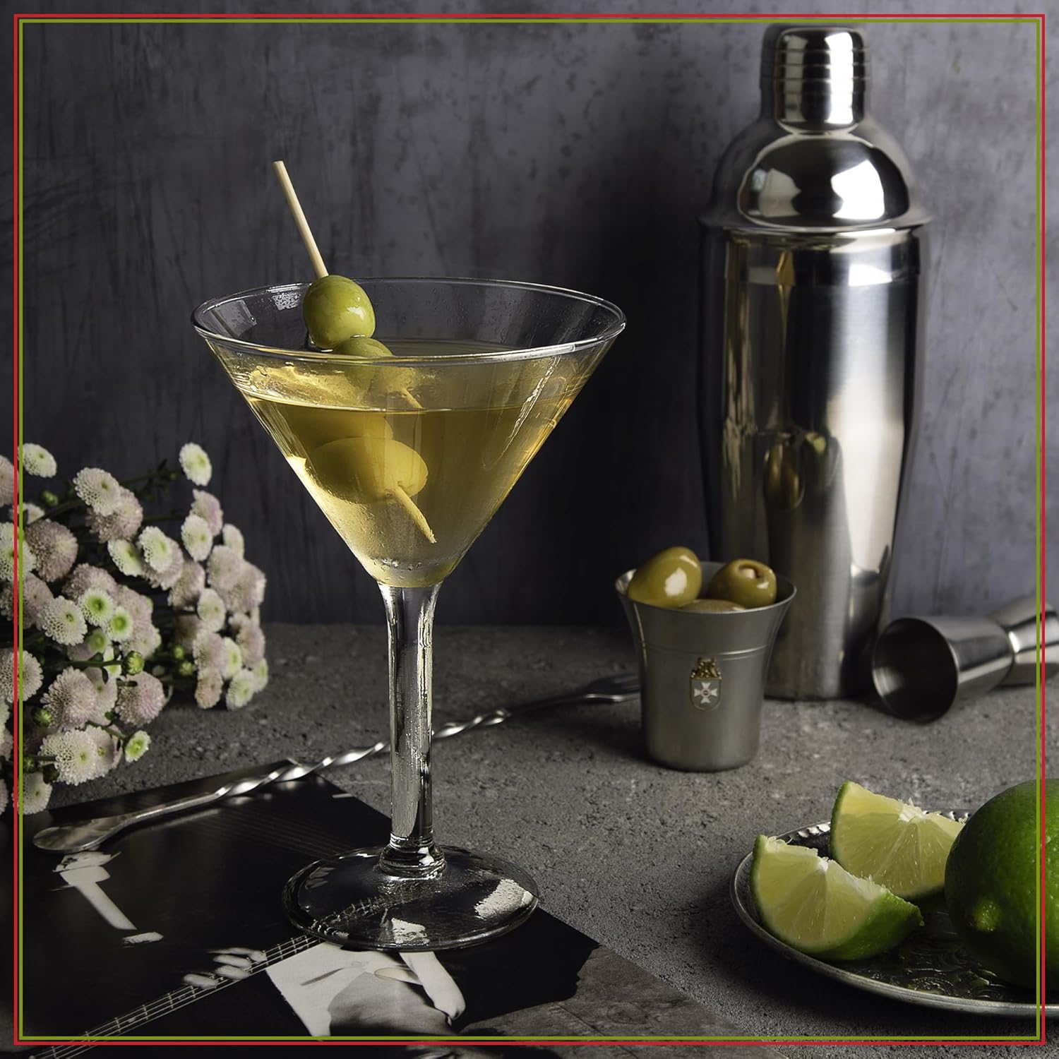 Cocktail with olives in a martini glass on a dark surface with a cocktail shaker and limes.