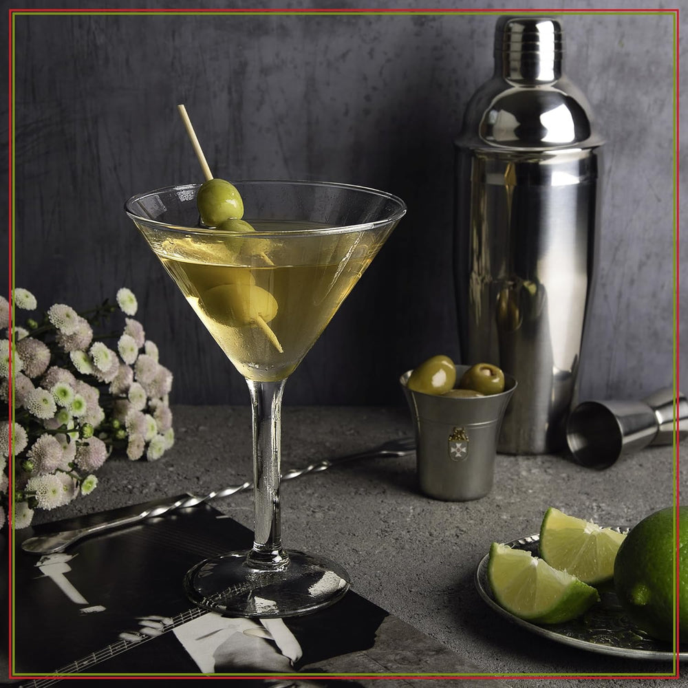 Cocktail with olives in a martini glass on a dark surface with a cocktail shaker and limes.