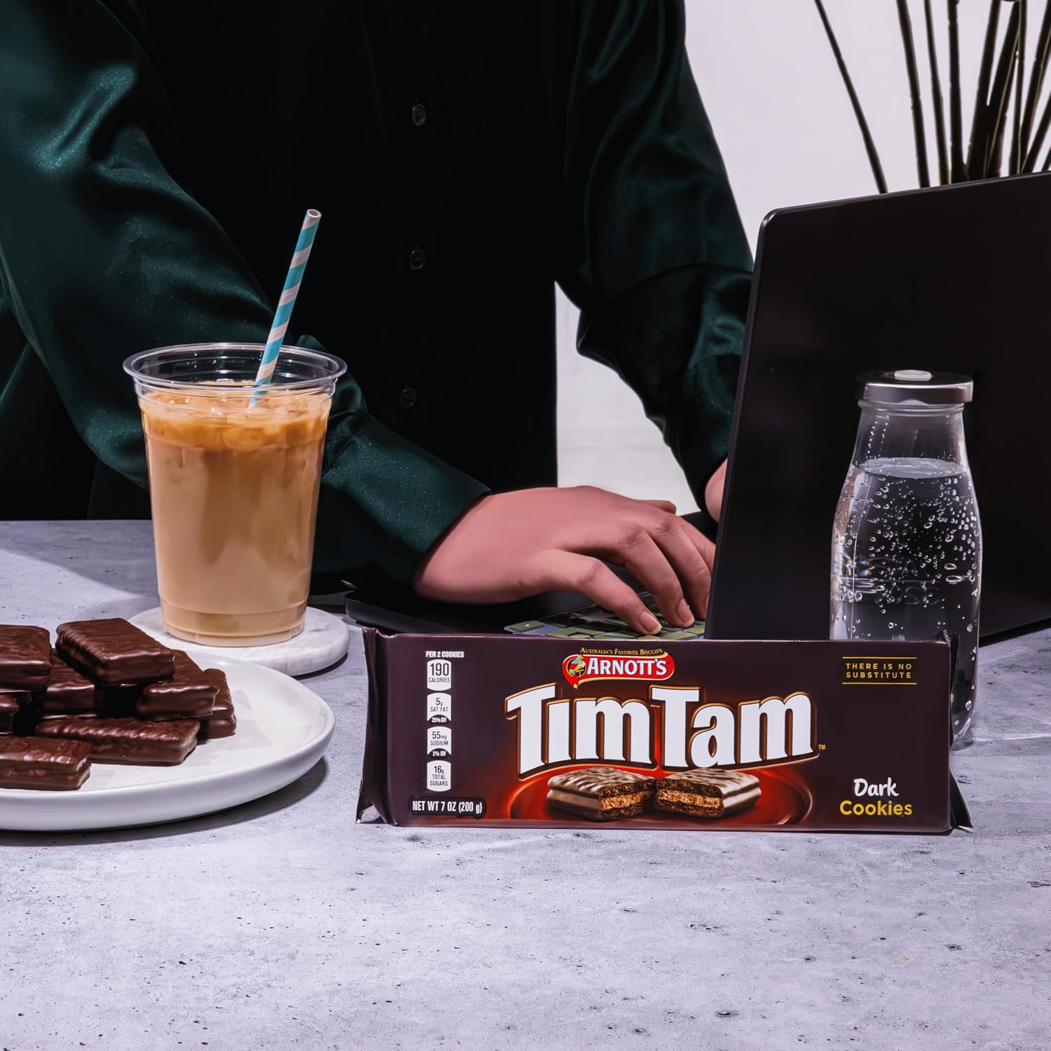 A person with a laptop enjoying dark chocolate Tim Tam cookies and iced coffee in a cozy setting.